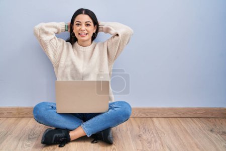 Foto de Mujer joven usando portátil sentado en el suelo en casa relajante y estiramiento, brazos y manos detrás de la cabeza y el cuello sonriendo feliz - Imagen libre de derechos