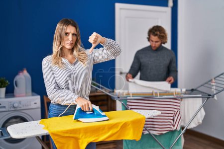 Photo for Couple ironing clothes at laundry room with angry face, negative sign showing dislike with thumbs down, rejection concept - Royalty Free Image