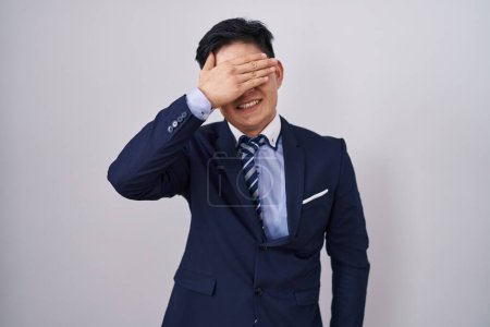 Téléchargez les photos : Jeune homme asiatique portant un costume d'affaires et cravate souriant et riant avec la main sur le visage couvrant les yeux pour la surprise. concept aveugle. - en image libre de droit