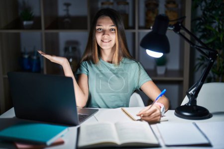 Photo for Teenager girl doing homework at home late at night smiling cheerful presenting and pointing with palm of hand looking at the camera. - Royalty Free Image