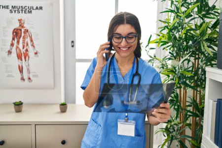 Foto de Mujer hispana joven vistiendo uniforme médico hablando en el teléfono inteligente buscando rayos X en la clínica - Imagen libre de derechos