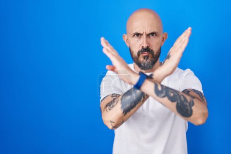 Foto de Hombre hispano con tatuajes de pie sobre fondo azul expresión de rechazo cruzando brazos haciendo signo negativo, cara enojada - Imagen libre de derechos