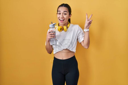 Photo for Young south asian woman wearing sportswear drinking water smiling with happy face winking at the camera doing victory sign with fingers. number two. - Royalty Free Image