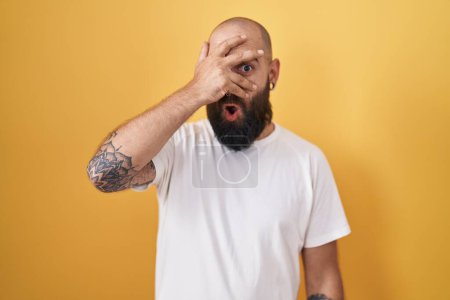 Photo for Young hispanic man with beard and tattoos standing over yellow background peeking in shock covering face and eyes with hand, looking through fingers with embarrassed expression. - Royalty Free Image