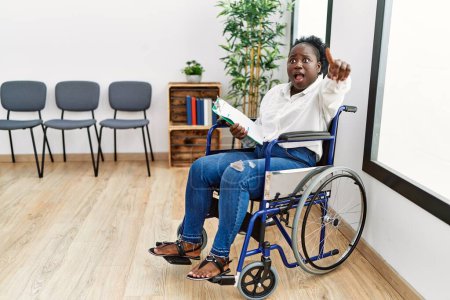 Téléchargez les photos : Jeune femme noire assise en fauteuil roulant dans la salle d'attente pointant du doigt surpris devant, bouche ouverte expression étonnante, quelque chose sur le devant - en image libre de droit
