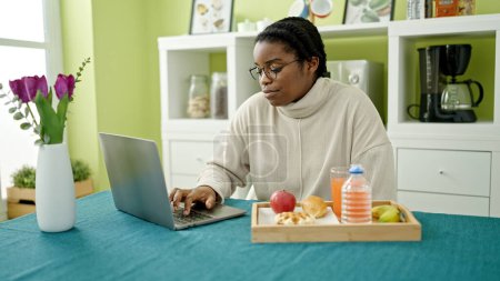 Téléchargez les photos : Femme afro-américaine prenant le petit déjeuner en utilisant un ordinateur portable à la salle à manger - en image libre de droit