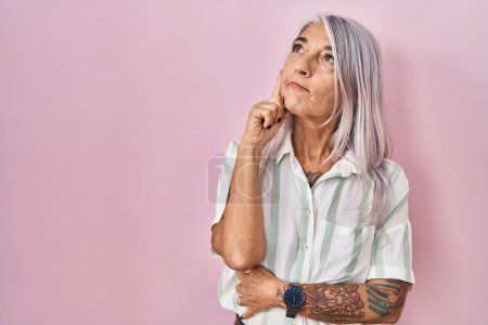 Téléchargez les photos : Femme d'âge moyen avec les cheveux gris debout sur fond rose visage sérieux penser à la question avec la main sur le menton, réfléchi sur idée déroutante - en image libre de droit