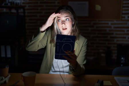 Photo for Blonde caucasian woman working at the office at night very happy and smiling looking far away with hand over head. searching concept. - Royalty Free Image