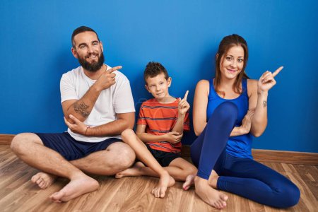 Photo for Family of three sitting on the floor at home with a big smile on face, pointing with hand and finger to the side looking at the camera. - Royalty Free Image