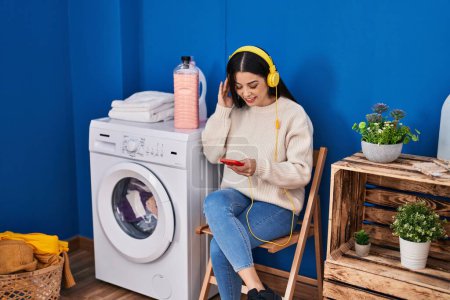 Photo for Young beautiful hispanic woman listening to music waiting for washing machine at laundry room - Royalty Free Image