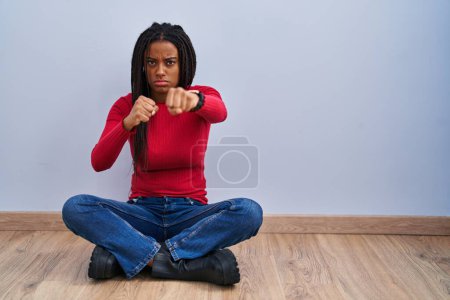 Foto de Joven afroamericano con trenzas sentado en el suelo en casa golpeando puño para luchar, ataque agresivo y enojado, amenaza y violencia - Imagen libre de derechos