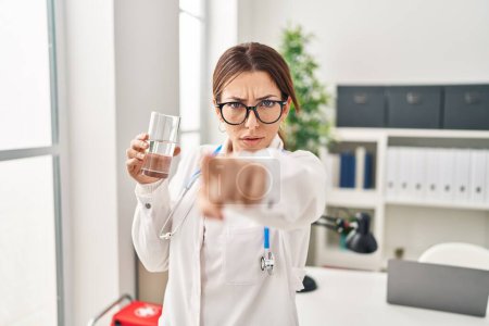 Téléchargez les photos : Jeune femme brune médecin tenant un verre d'eau pointant du doigt vers la caméra et vers vous, geste confiant et sérieux - en image libre de droit
