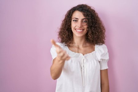 Foto de Mujer hispana con el pelo rizado de pie sobre fondo rosa sonriendo amistoso ofreciendo apretón de manos como saludo y bienvenida. negocios exitosos. - Imagen libre de derechos