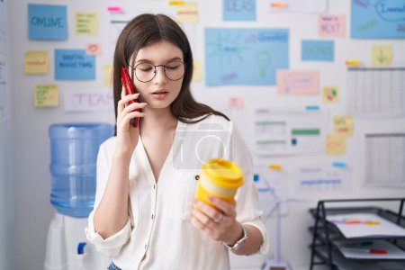 Téléchargez les photos : Young caucasian woman business worker talking on smartphone drinking coffee at office - en image libre de droit
