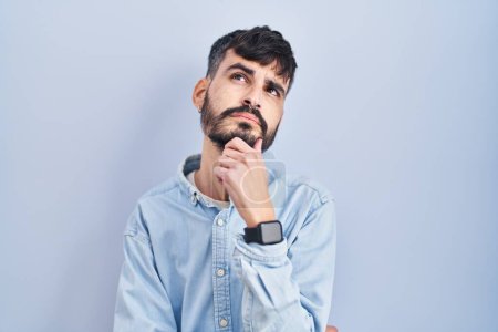 Photo for Young hispanic man with beard standing over blue background with hand on chin thinking about question, pensive expression. smiling with thoughtful face. doubt concept. - Royalty Free Image