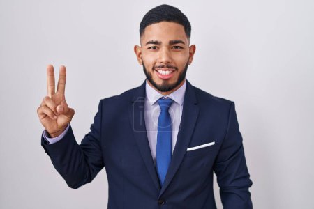 Téléchargez les photos : Jeune homme hispanique portant un costume d'affaires et une cravate souriant avec un clin d'œil heureux à la caméra faisant signe de victoire avec les doigts. numéro deux. - en image libre de droit