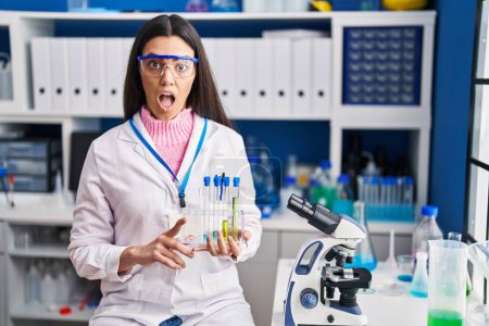 Téléchargez les photos : Jeune femme brune travaillant au laboratoire scientifique effrayée et étonnée avec la bouche ouverte pour surprise, visage d'incrédulité - en image libre de droit