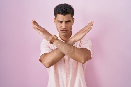 Foto de Joven hombre hispano de pie sobre fondo rosa expresión de rechazo cruzando brazos haciendo signo negativo, cara enojada - Imagen libre de derechos