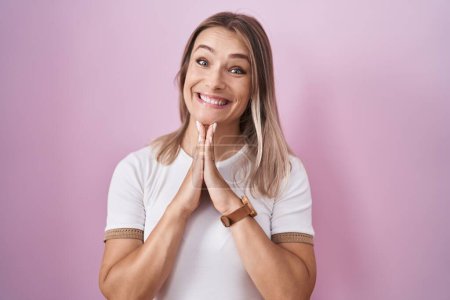 Foto de Blonde caucasian woman standing over pink background praying with hands together asking for forgiveness smiling confident. - Imagen libre de derechos