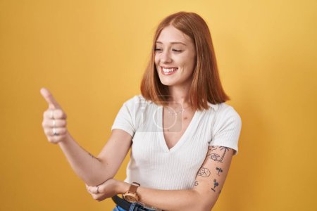 Téléchargez les photos : Jeune rousse femme debout sur fond jaune regardant fier, souriant faire les pouces geste vers le haut sur le côté - en image libre de droit
