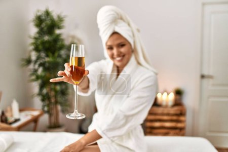 Foto de Joven mujer hispana hermosa usando albornoz bebiendo champán en el salón de belleza - Imagen libre de derechos