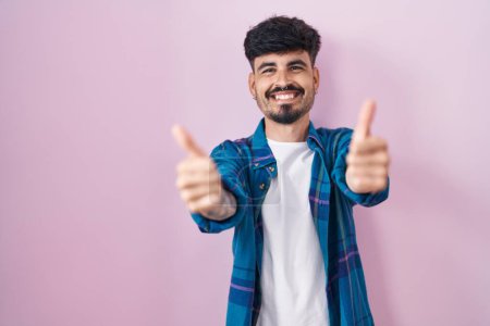 Téléchargez les photos : Jeune homme hispanique avec la barbe debout sur fond rose approuvant faire un geste positif avec la main, pouces levés souriant et heureux pour le succès. geste gagnant. - en image libre de droit