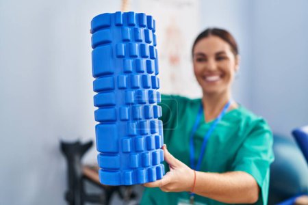 Téléchargez les photos : Young beautiful hispanic woman physiotherapist smiling confident holding foam roller at rehab clinic - en image libre de droit