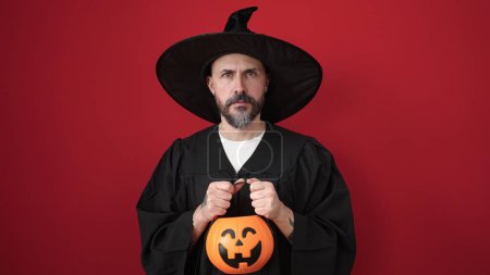 Photo for Young bald man wearing wizard costume holding pumpkin halloween basket over isolated red background - Royalty Free Image