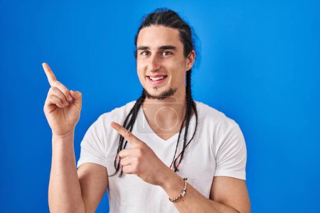 Téléchargez les photos : Homme hispanique aux cheveux longs debout sur fond bleu souriant et regardant la caméra pointant avec deux mains et doigts sur le côté. - en image libre de droit