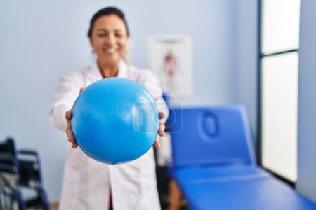 Photo for Middle age hispanic woman physiotherapist holding ball at rehab clinic - Royalty Free Image