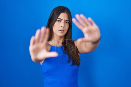 Foto de Mujer hispana de pie sobre fondo azul haciendo marco usando manos palmas y dedos, perspectiva de la cámara - Imagen libre de derechos