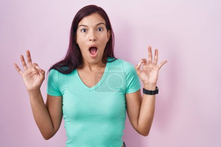 Photo for Young hispanic woman standing over pink background looking surprised and shocked doing ok approval symbol with fingers. crazy expression - Royalty Free Image