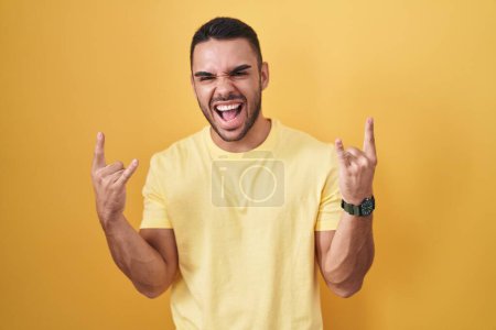 Téléchargez les photos : Jeune homme hispanique debout sur fond jaune criant avec une expression folle faire symbole rock avec les mains en l'air. star de la musique. concept de musique lourde. - en image libre de droit