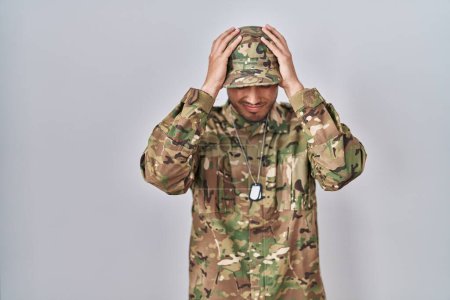Foto de Joven hispano vestido con uniforme camuflaje del ejército que sufre de dolor de cabeza desesperado y estresado debido al dolor y la migraña. manos en la cabeza. - Imagen libre de derechos
