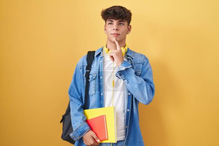 Téléchargez les photos : Adolescent hispanique portant un sac à dos d'étudiant et tenant des livres pensant concentré sur le doute avec le doigt sur le menton et regardant vers le haut se demandant - en image libre de droit