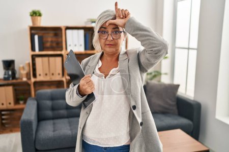 Photo for Middle age woman with grey hair at consultation office making fun of people with fingers on forehead doing loser gesture mocking and insulting. - Royalty Free Image