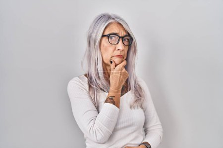 Téléchargez les photos : Femme d'âge moyen avec les cheveux gris debout sur fond blanc pensant inquiet d'une question, préoccupé et nerveux avec la main sur le menton - en image libre de droit