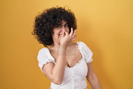 Téléchargez les photos : Jeune femme brune aux cheveux bouclés debout sur fond jaune sentant quelque chose de puant et dégoûtant, odeur intolérable, retenant son souffle avec les doigts sur le nez. mauvaise odeur - en image libre de droit