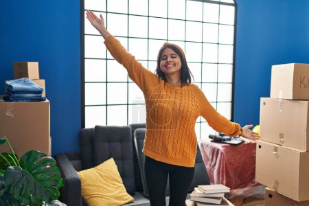 Téléchargez les photos : Jeune femme latine souriante confiante debout avec les bras ouverts à la nouvelle maison - en image libre de droit