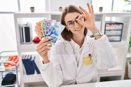 Téléchargez les photos : Jeune médecin caucasien femme tenant des billets de banque en dollars canadiens souriant heureux faisant ok signe avec la main sur les yeux regardant à travers les doigts - en image libre de droit