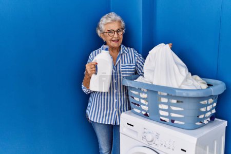 Photo for Senior grey-haired woman smiling confident holding detergent bottle at laundry room - Royalty Free Image