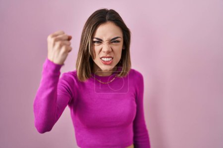 Foto de Mujer hispana de pie sobre fondo rosa enojado y loco levantando el puño frustrado y furioso mientras gritaba con ira. rabia y concepto agresivo. - Imagen libre de derechos