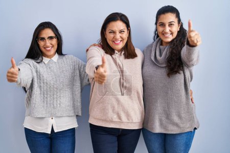 Photo for Mother and two daughters standing over blue background approving doing positive gesture with hand, thumbs up smiling and happy for success. winner gesture. - Royalty Free Image