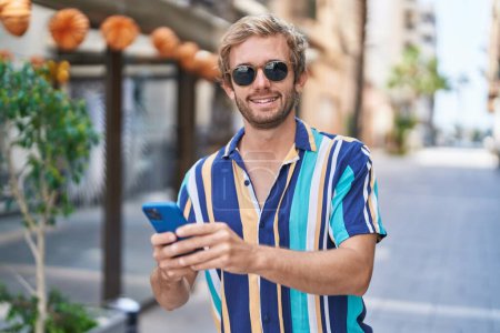 Téléchargez les photos : Jeune homme touriste souriant confiant en utilisant smartphone à la rue - en image libre de droit