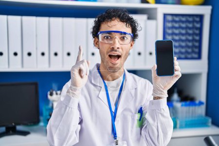 Foto de Hombre hispano trabajando en laboratorio científico mostrando pantalla de teléfono inteligente sorprendido con una idea o pregunta apuntando con el dedo con la cara feliz, número uno - Imagen libre de derechos