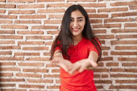 Téléchargez les photos : Jeune adolescente debout sur un mur de briques souriant avec les mains paumes ensemble recevoir ou donner geste. maintien et protection - en image libre de droit