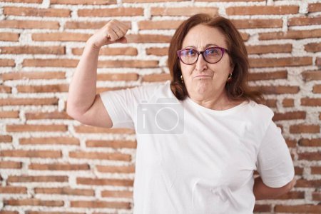 Téléchargez les photos : Femme âgée avec des lunettes debout sur le mur de briques personne forte montrant muscle du bras, confiant et fier de la puissance - en image libre de droit