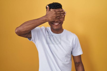 Foto de Young hispanic man standing over yellow background smiling and laughing with hand on face covering eyes for surprise. blind concept. - Imagen libre de derechos
