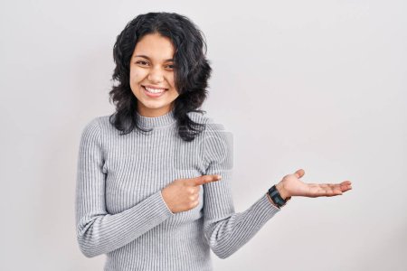 Photo for Hispanic woman with dark hair standing over isolated background amazed and smiling to the camera while presenting with hand and pointing with finger. - Royalty Free Image