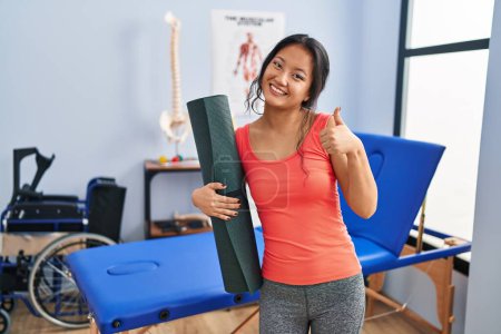 Foto de Joven asiática sosteniendo yoga mat en clínica sonriendo feliz y positiva, pulgar hacia arriba haciendo excelente y aprobación signo - Imagen libre de derechos